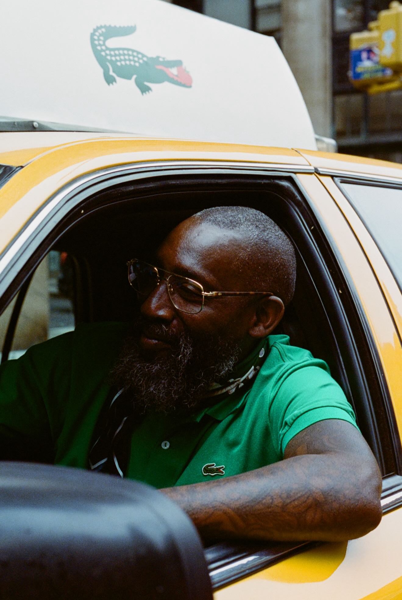 Taxi driver seen wearing a green Lacoste polo shirt leans out of a yellow taxi window on New York street.
