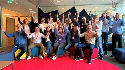 A group of people cheerfully posing together in a brightly lit room, with a red carpet and playful expressions.