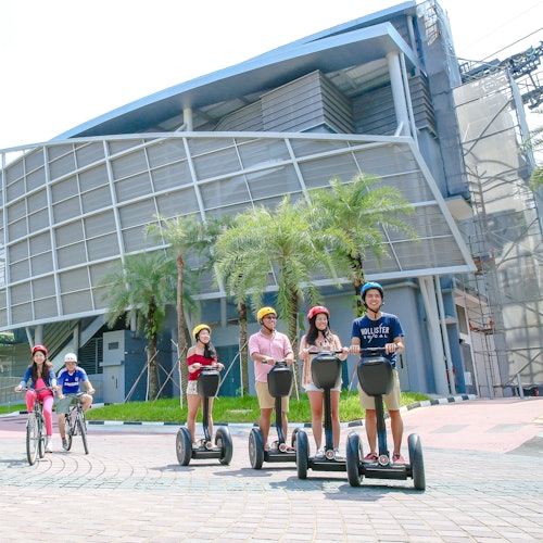 Group of people riding Segways and bicycles in front of a modern building with palm trees and a tiled walkway.