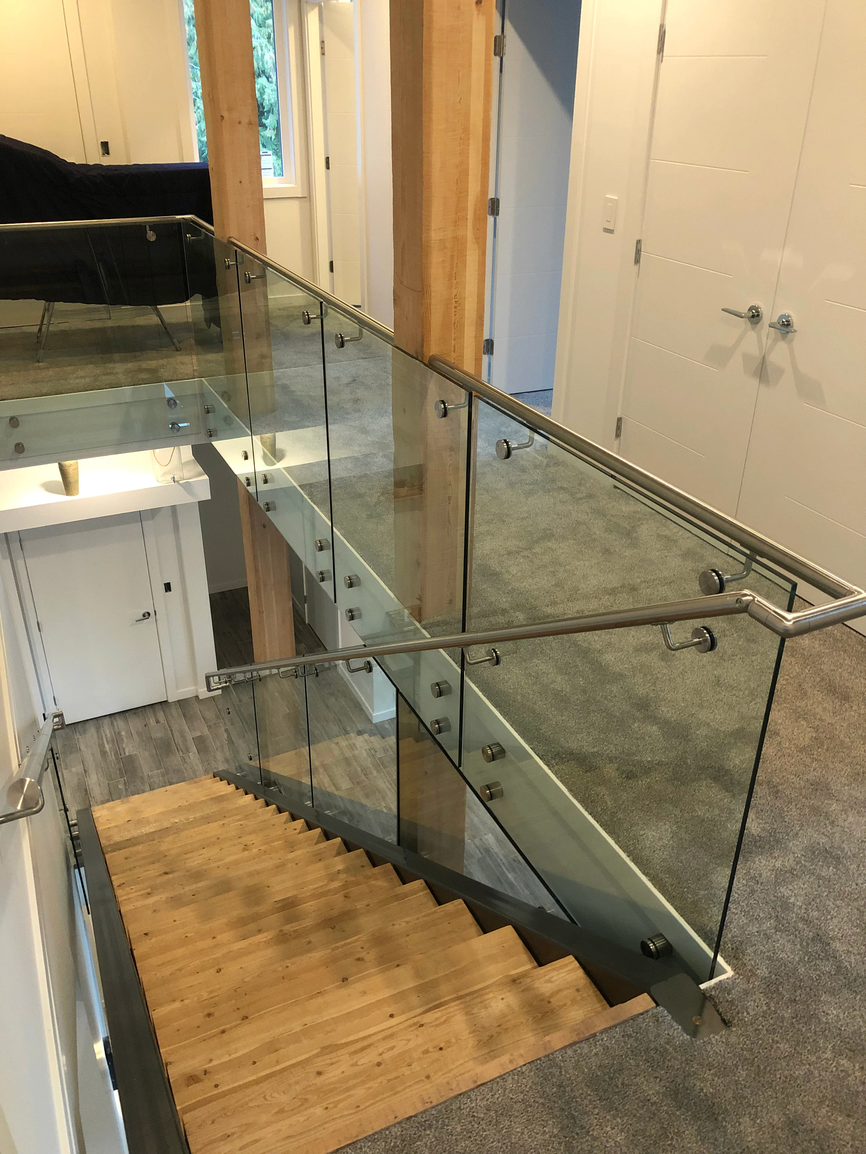 An interior loft railing with glass panels mounted to a dark structural frame using paired round stainless steel standoffs, topped with a brushed stainless tubular handrail. Wood support beams and carpeted flooring surround the opening, with a wood staircase leading down to a lower level and closet doors visible along the hallway.