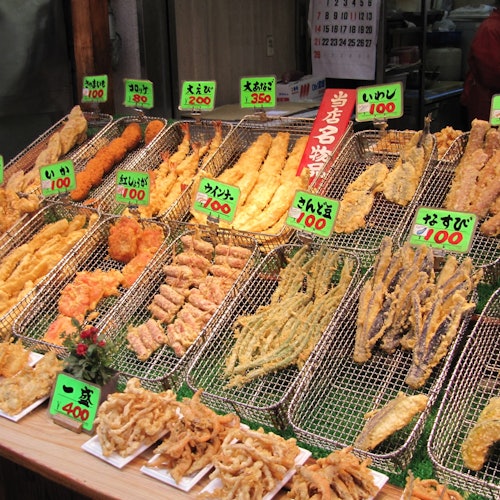 展示各种炸食品的陈列架上挂着色彩鲜艳的价格标签，用日文标识，可能是在市场或食品摊位上的地方。