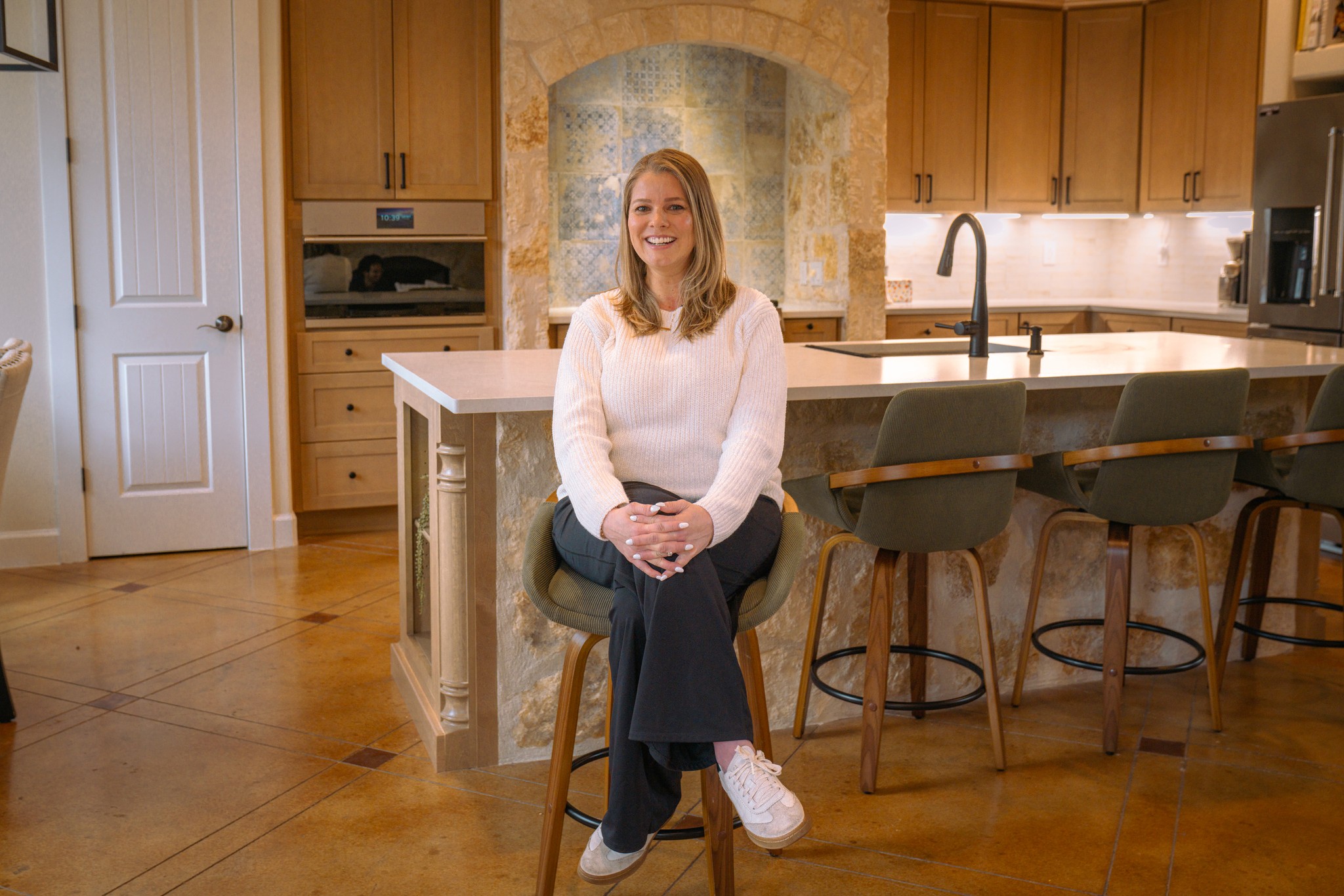Happy QW Kitchen & Bath client seated at custom kitchen island with natural oak cabinets and stone backsplash in Austin Texas