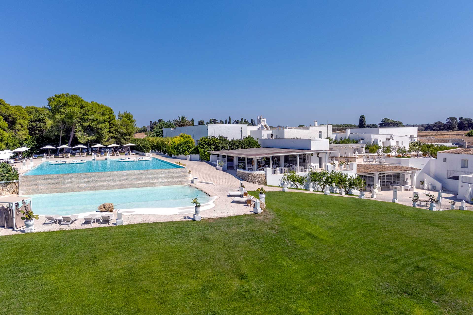 Luftaufnahme eines Hotels mit Swimmingpool, Rasenfläche und weißen Gebäuden unter blauem Himmel.