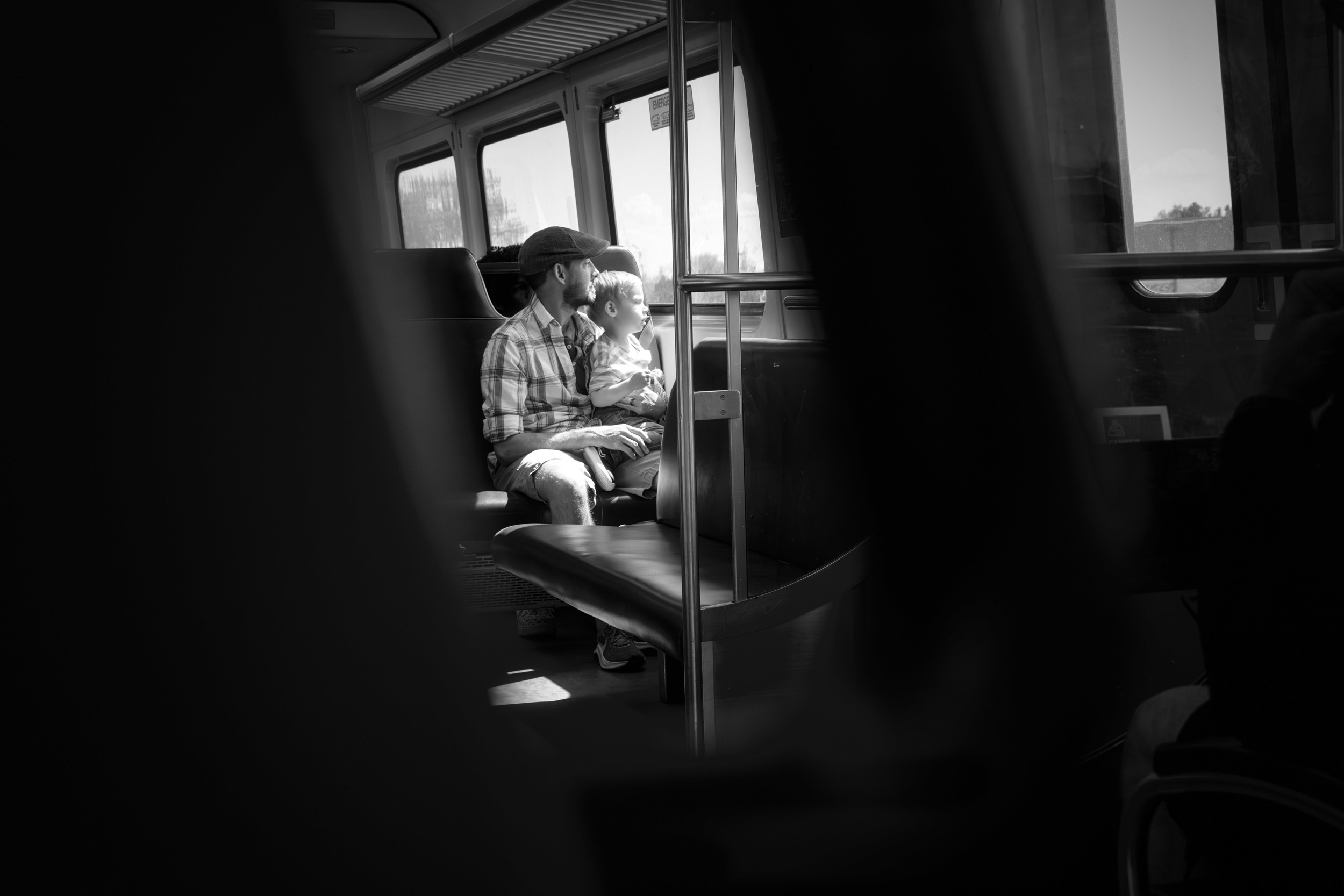 A man and child sit together on a bus, bathed in soft window light, both gazing outside with wonder. Photographed through dark foreground shapes that create a natural frame within the frame.