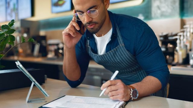 Em ambiente de cafeteria, homem pardo segura uma caneta sobre prancheta em balcão com tablet em suporte, enquanto está ao celular.