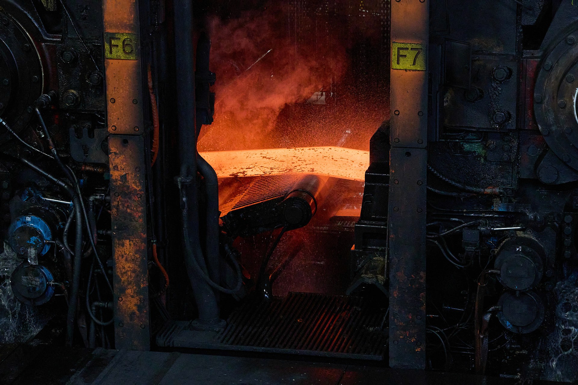 Gros plan sur une plaque d'acier en fusion rouge vif sortant d'un laminoir industriel, générant de la fumée et des projections d'eau.