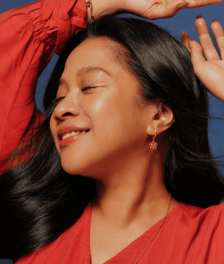 A joyful woman with long black hair, smiling while wearing a red top and gold earrings, with her hands raised against a blue background.