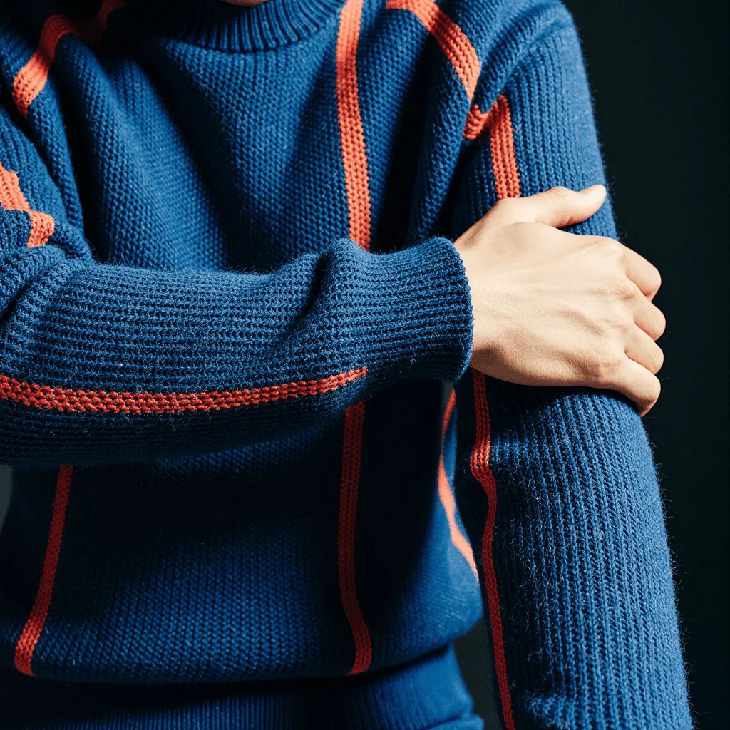 Une personne portant un pull en maille bleu avec des rayures verticales orange sur les manches et le corps, son bras croisé sur la poitrine, mettant en valeur la texture du tricot.