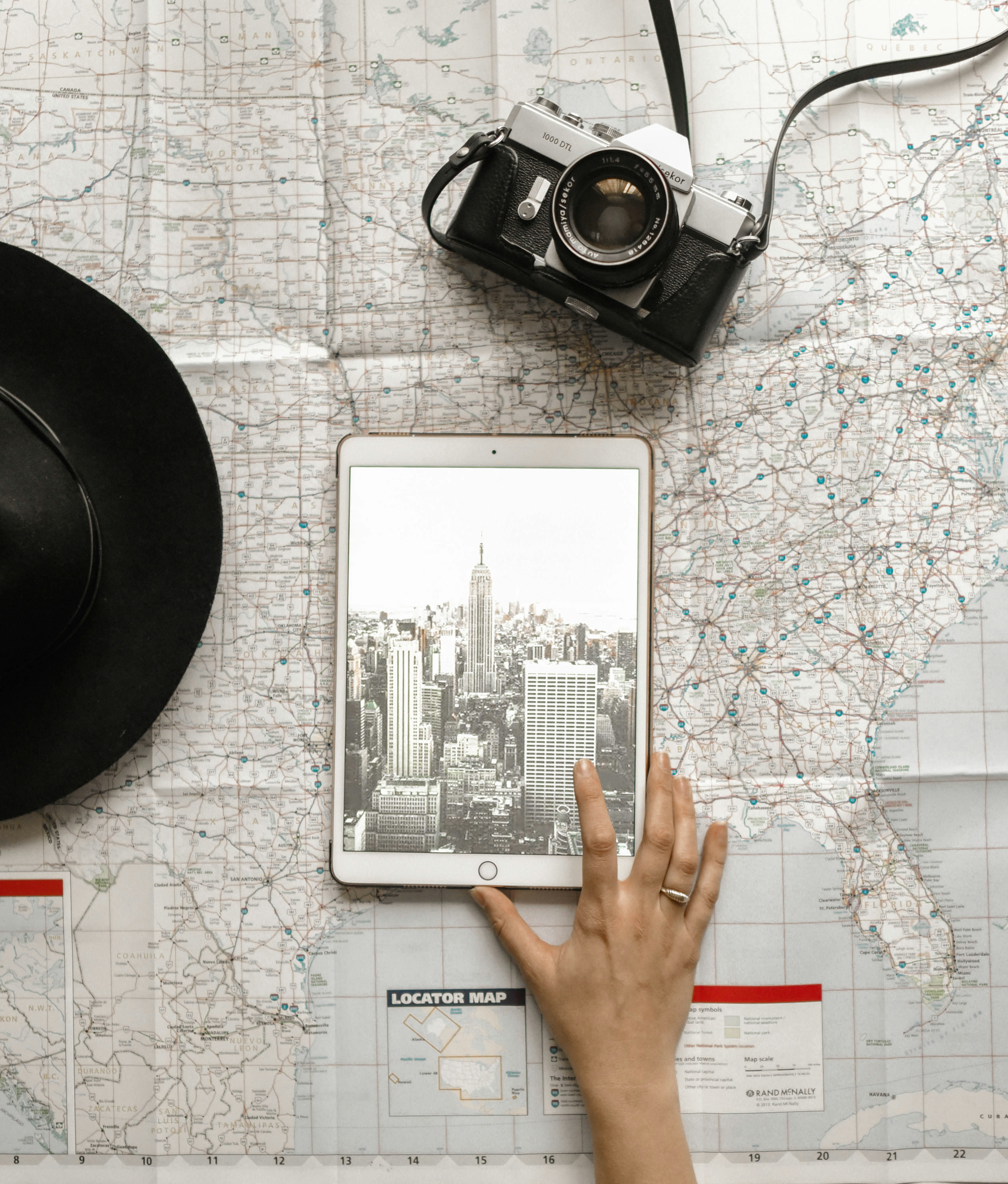 An overhead view of a traveler's planning table with a map spread out, showing a hand resting on a tablet displaying a black and white photo of a cityscape, likely for trip inspiration. A classic film camera and a stylish hat suggest a journey full of adventure and photography.