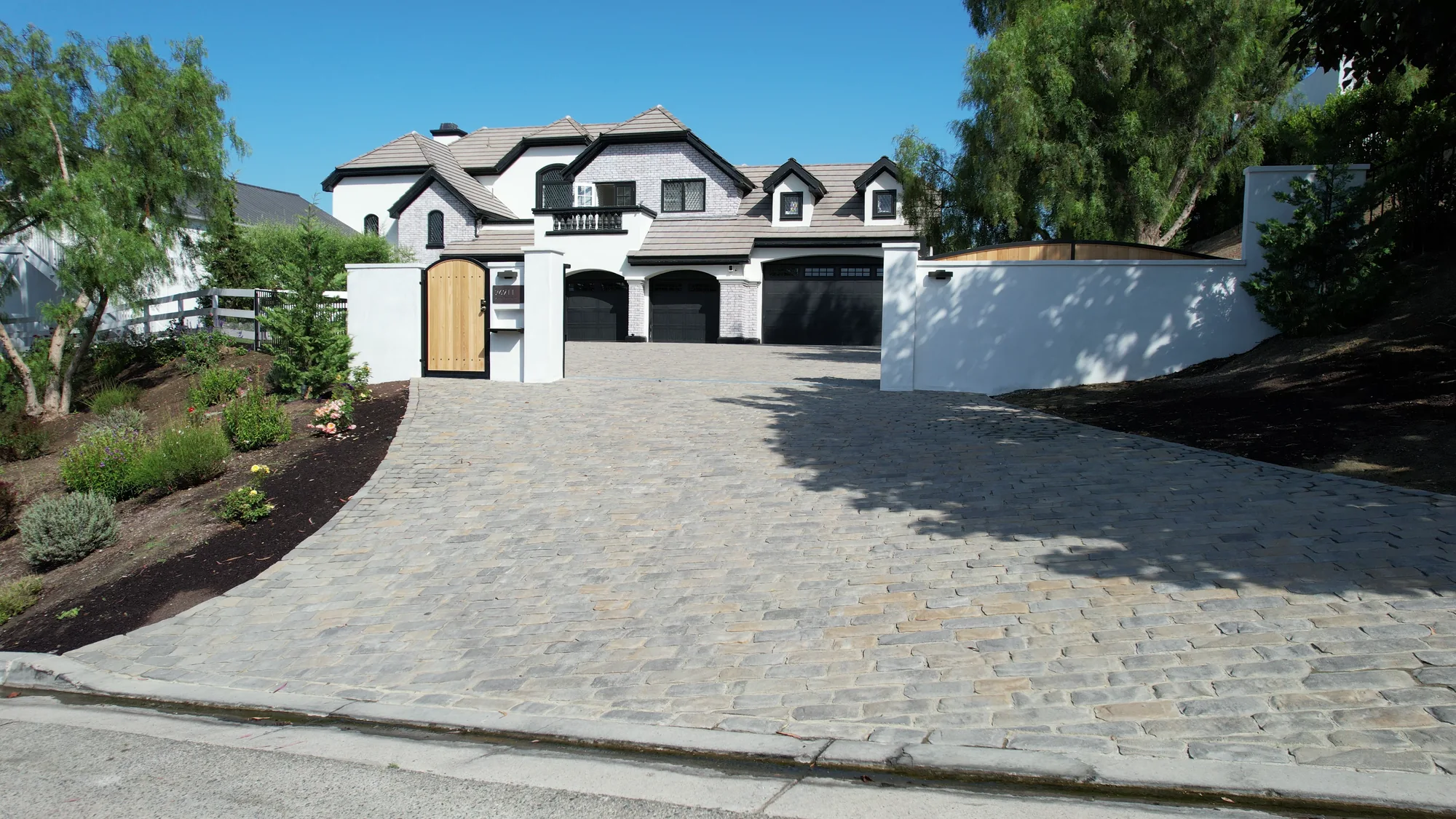 Daylight view up a broad gray paver driveway toward open gates and a white two-story home.