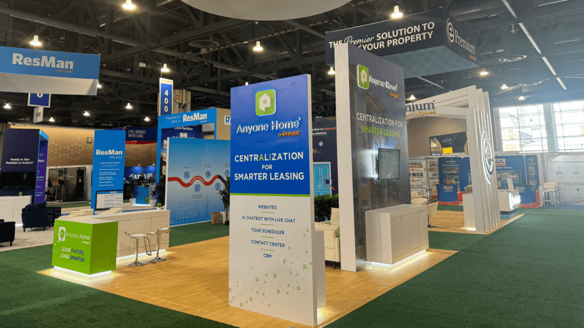 Anyone Home 20x20 professional service booth at NAA 2024 showing overhead circular hanging sign, green brand tower, demo screen, reception counter, and aisle-facing booth visibility.