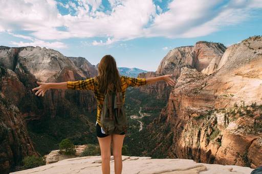 A person standing on a cliff edge with arms outstretched, overlooking a beautiful canyon landscape.