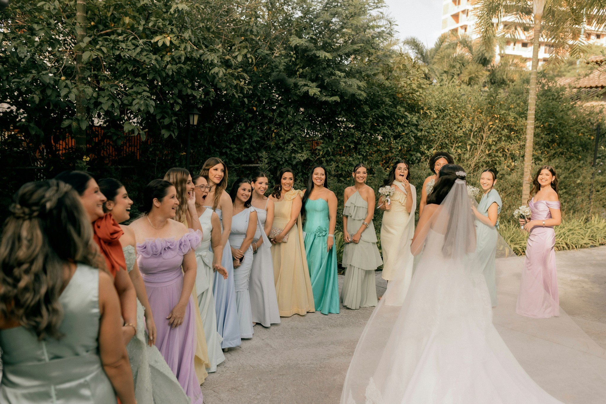 Bridesmaids in colorful pastel dresses reacting with joy as the bride reveals her wedding gown in an outdoor first look