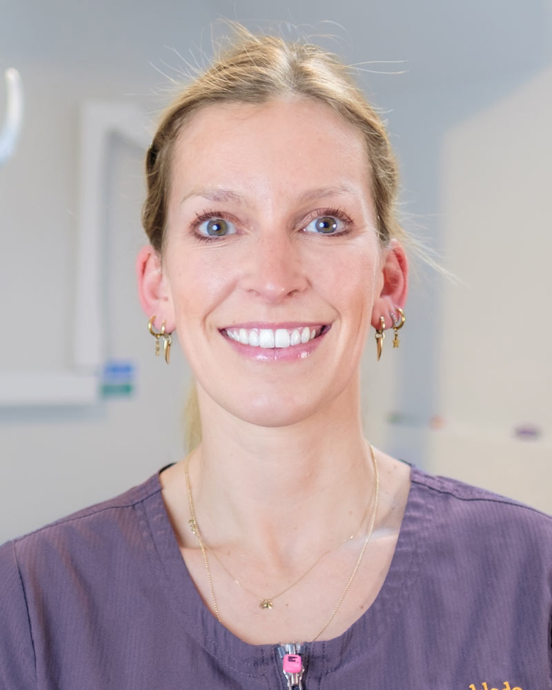 A portrait of Julia, Principal Dentist at Cricklade Dental Practice, smiling and wearing a dark grey scrub top with a pink zipper pull and yellow embroidered text that reads "JULIA" on the left and "Cricklade Dental Practice" on the right. She is standing in a dental surgery with a dental light and other equipment visible in the background.