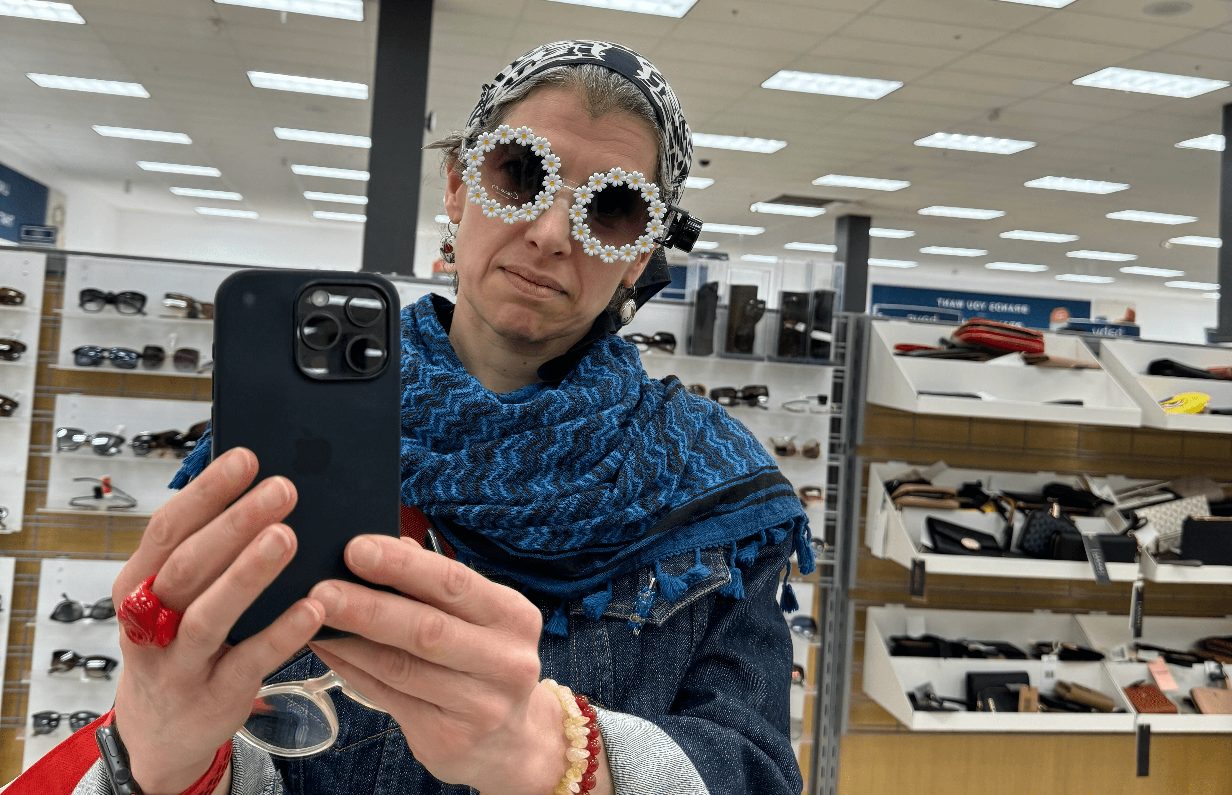 A lady wearing a scarf on her head and a blue keffiyeh taking a selfie in a department store of herself in a goofy pair of sunglasses whose frames are a ring of daisies. Photo by Rafia Mahli