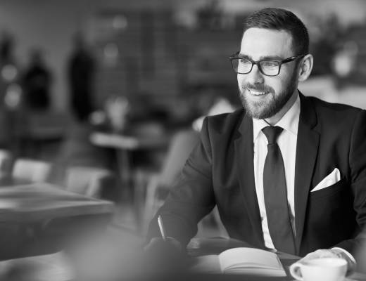 Smiling man in a suit and tie, wearing glasses, sitting at a table in a café or office space with a notebook and coffee cup in front of him.
