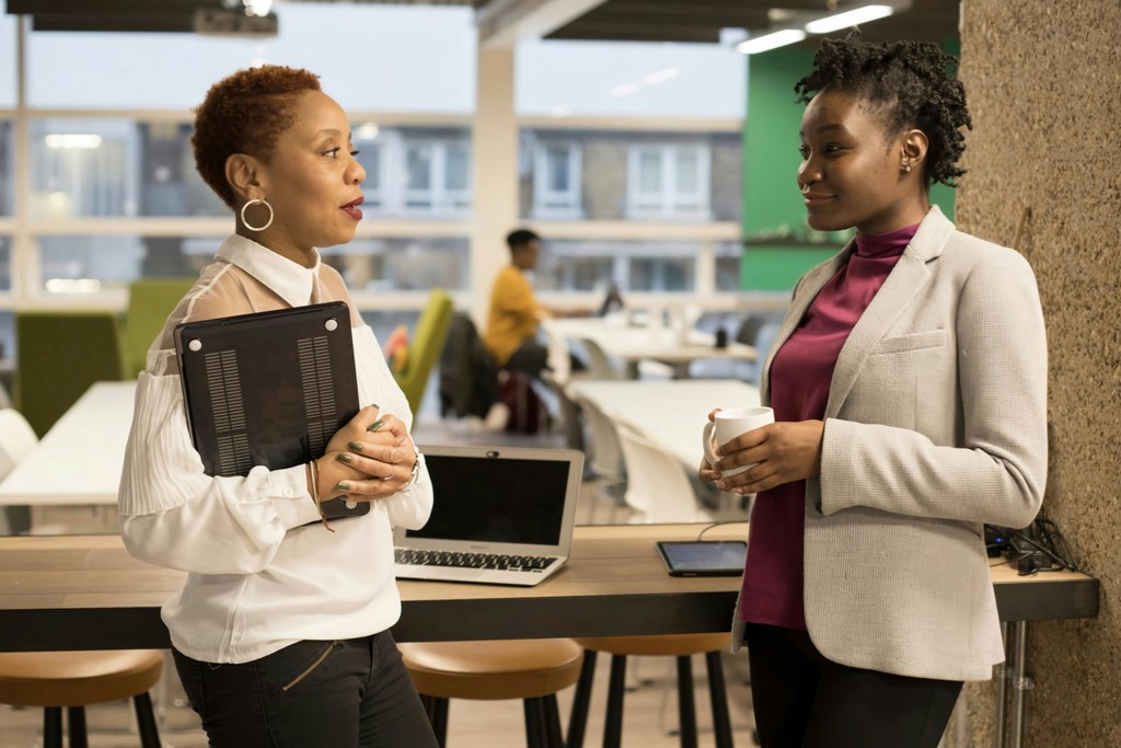 Two professional women engage in a conversation in a modern, open office space, with one holding a tablet and the other a coffee cup, highlighting collaboration and networking in the workplace.