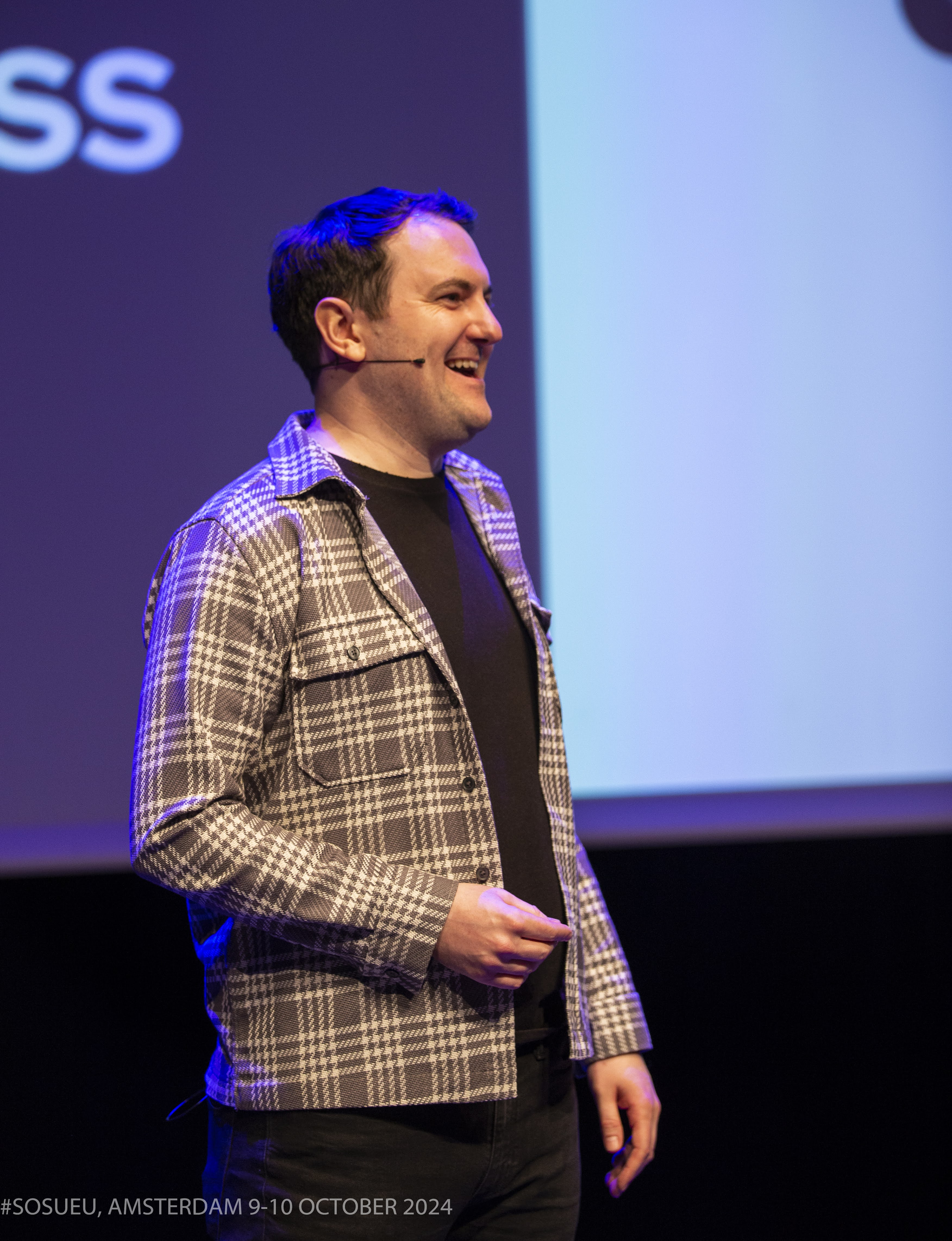 Matt Mercer stands on stage, wearing a plaid jacket, giving a presentation with a backdrop of text.
