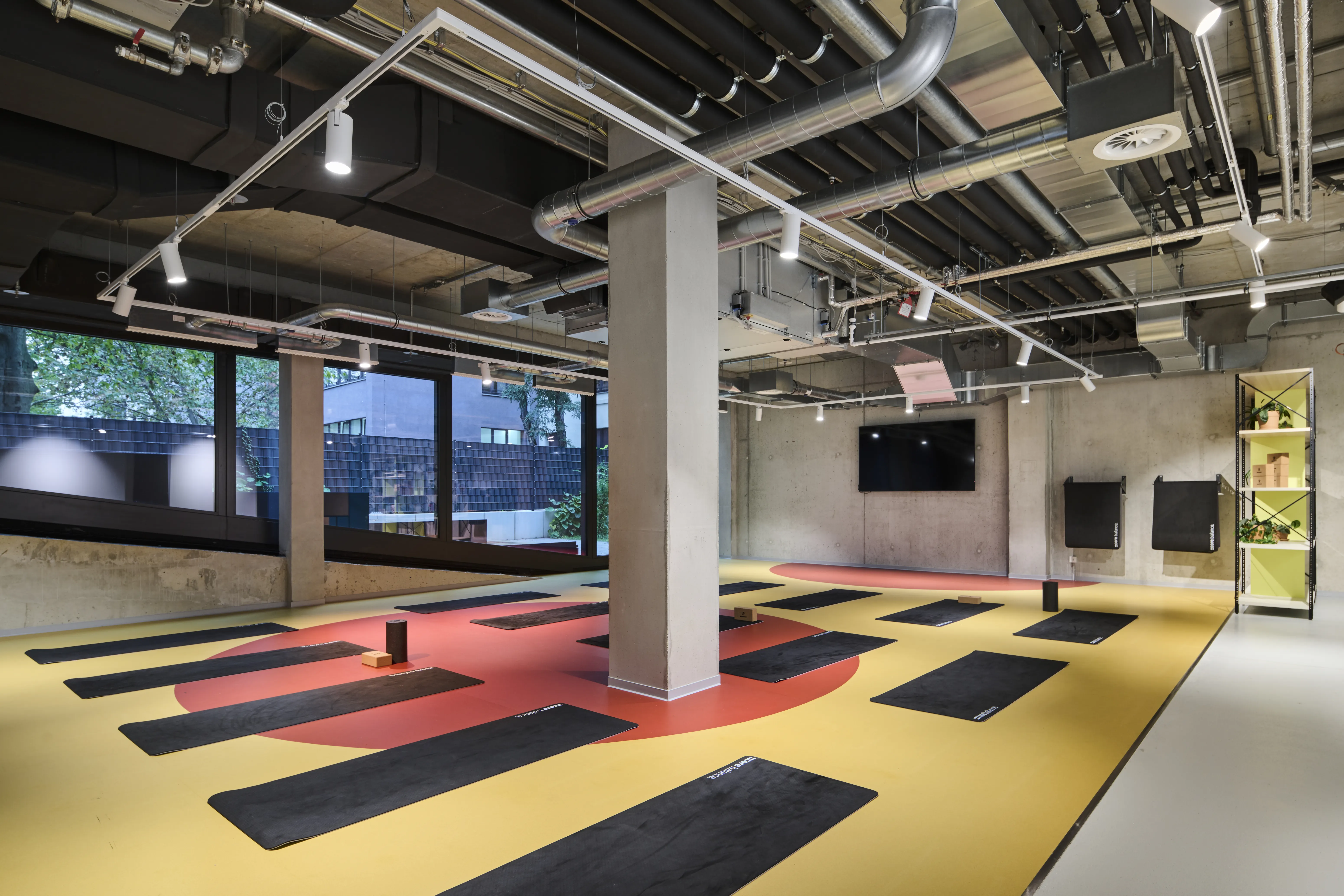 Course room at HELLOFRESH GYM with black yoga mats on a yellow floor with a large red circle, wall-mounted TV and window front with greenery.