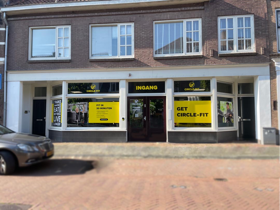 A Circle Fit hub with large windows displaying yellow signs, located on a brick street with a parked car nearby.