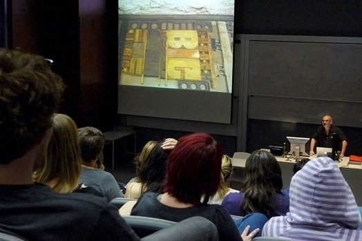 A group of people watching a projected screen video that shows metallic letters used on a printing press.
