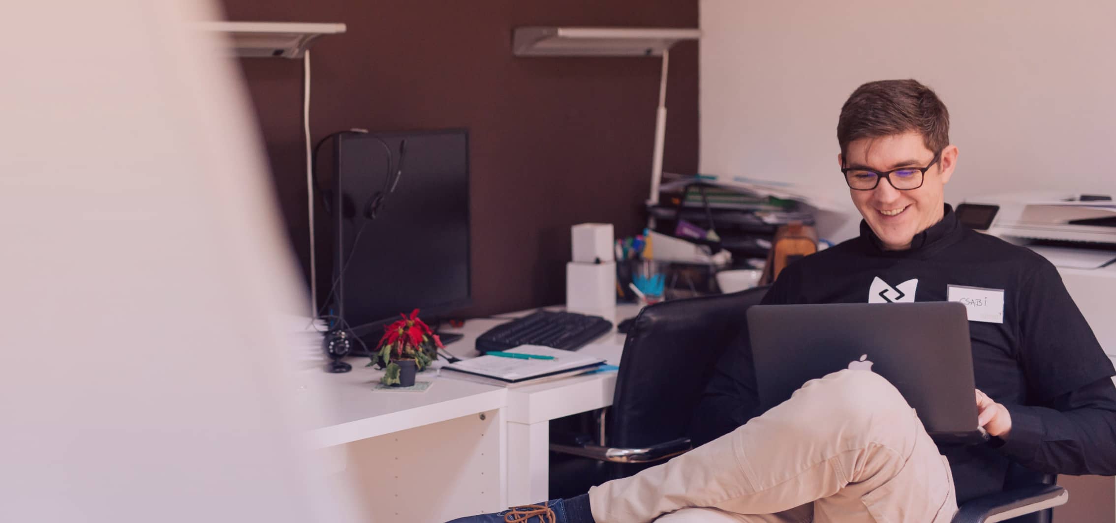 A photo of a man sitting on a chair and working on a laptop with a smile.