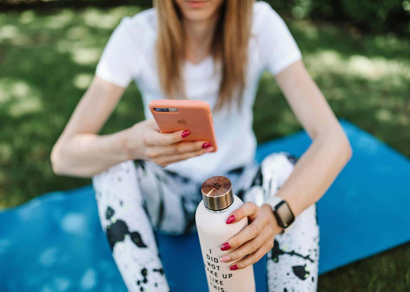 Woman sitting on blue yoga mat and holding a water bottle looking at iPhone