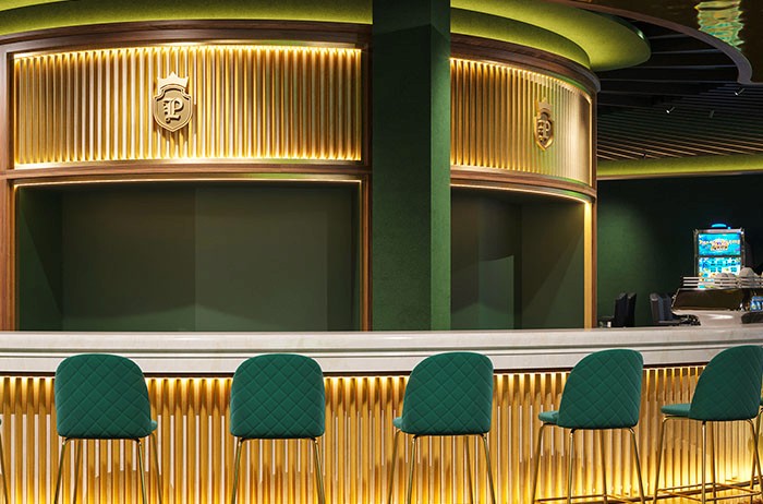 Innenansicht einer eleganten Bar mit goldenem Rippentresen, grünen gepolsterten Barhockern und Wappenverzierung – Gastronomiedesign