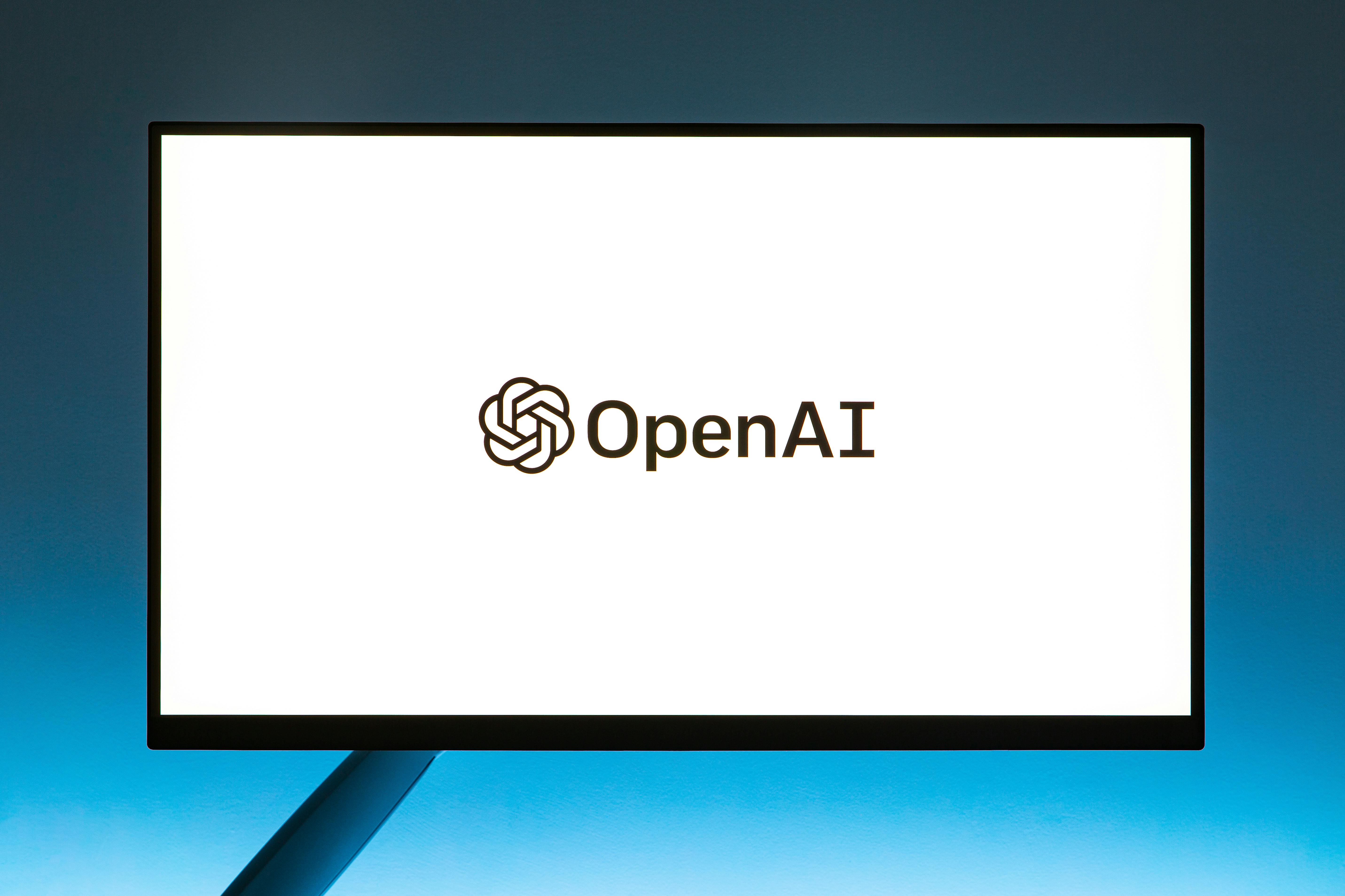 A computer screen displays the OpenAI logo, featuring a stylized neural node symbol and the word "OpenAI" in black, set against a white background.