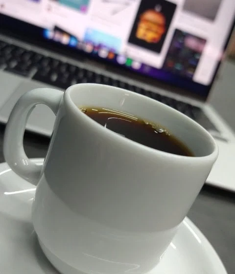 Uma xícara de café branca repousa sobre um pequeno prato com café escuro, em frente a um laptop que exibe diversas imagens.