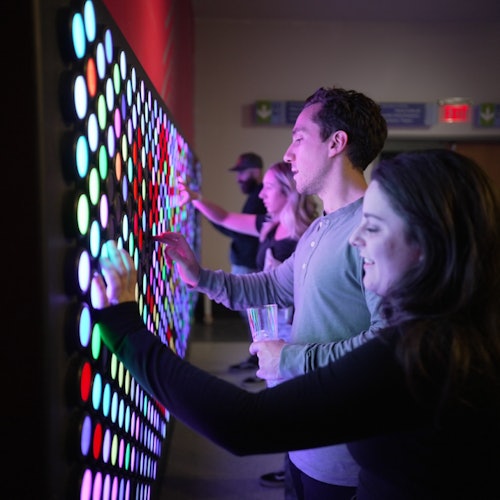 Las personas interactúan con una pared de botones coloridos e iluminados en una habitación con poca luz.
