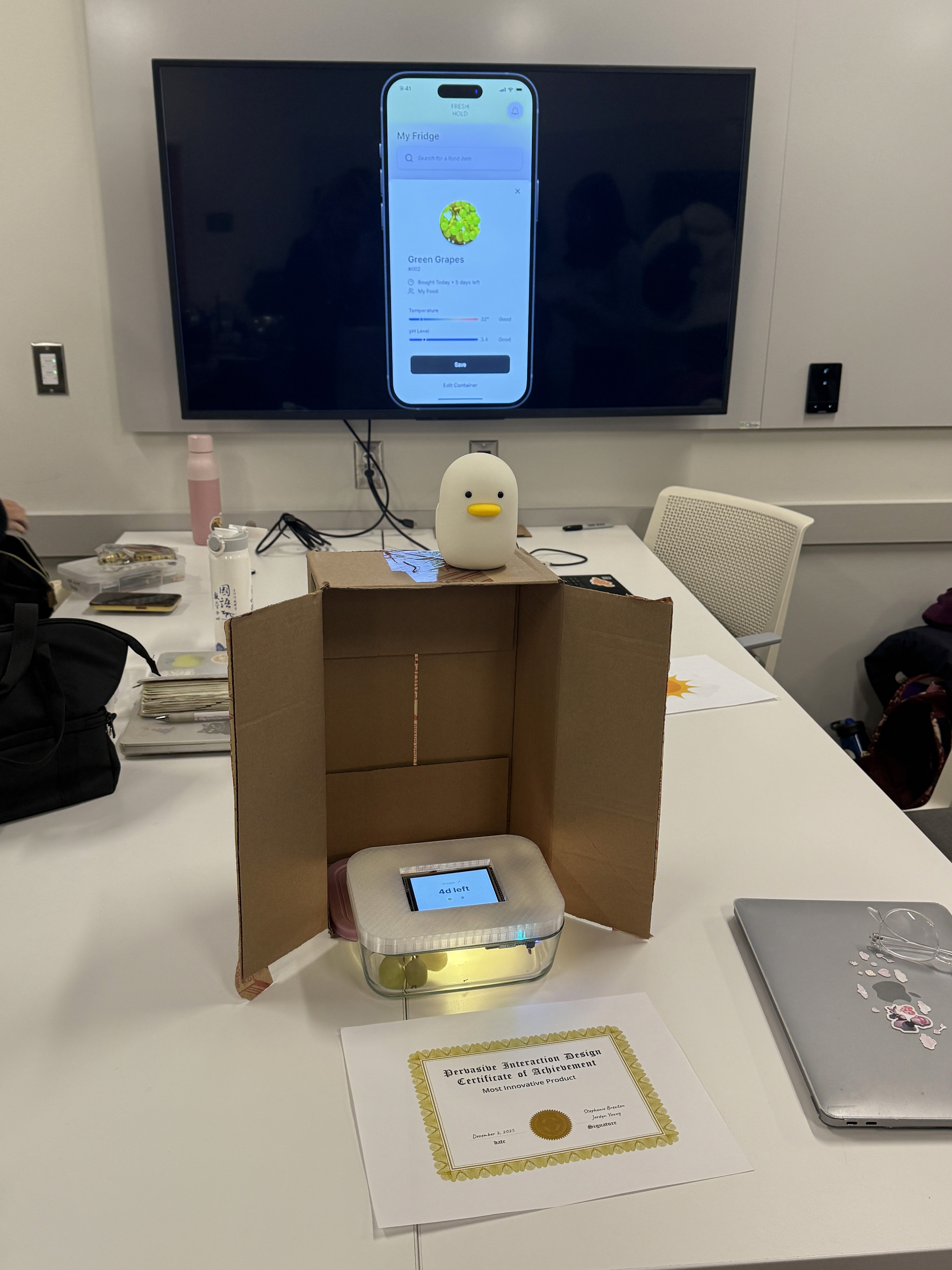 App prototype shown on a TV screen, with a makeshift cardboard fridge with the Freshhold container in it. A certificate sits on the table saying "Most Innovative Product."