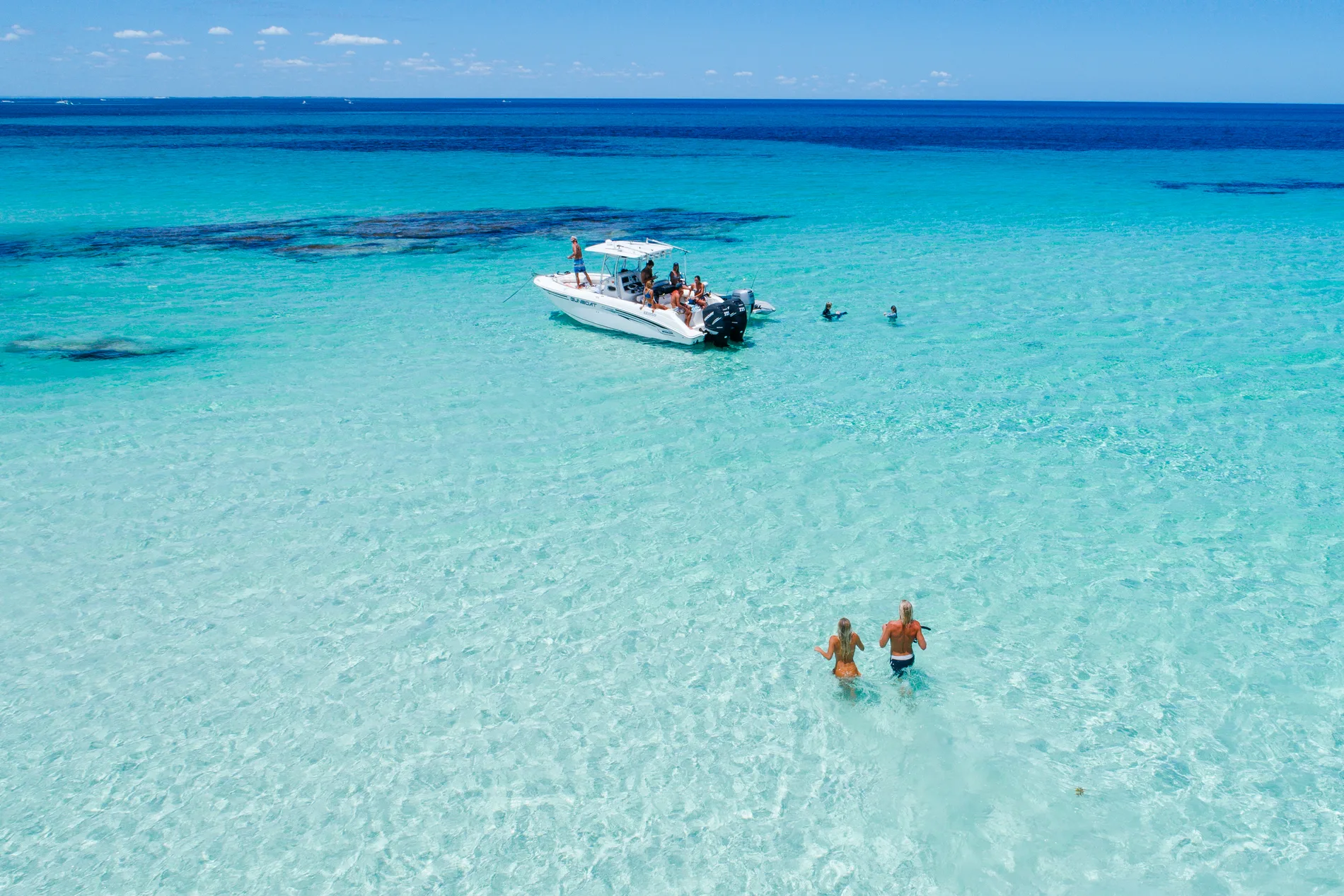 A boat anchored in shallow turquoise water with two people wading ashore
