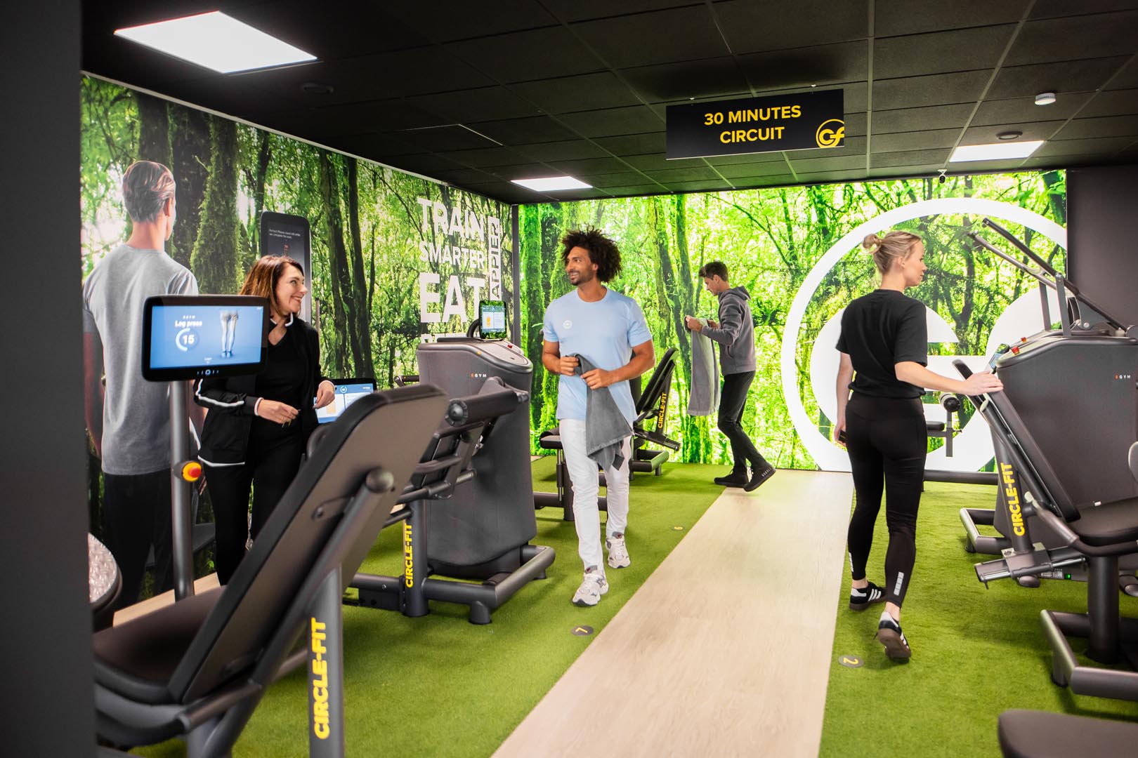 A modern gym interior with green accents, featuring people using fitness equipment and exercising in the Circle Fit circuit.