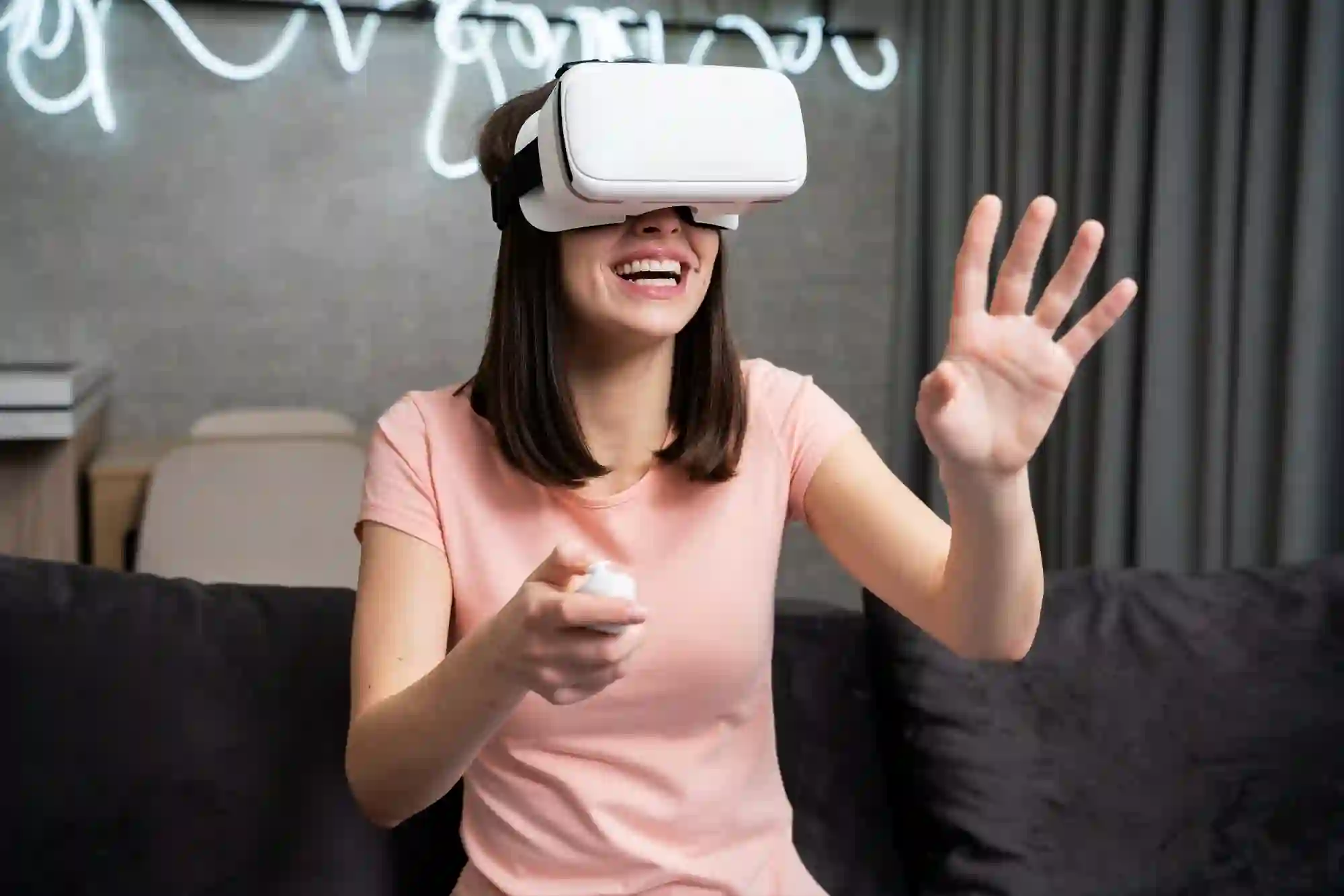 A smiling young woman wearing a white virtual reality (VR) headset sits on a couch, holding a controller and reaching out with her left hand, engaging with a virtual world.