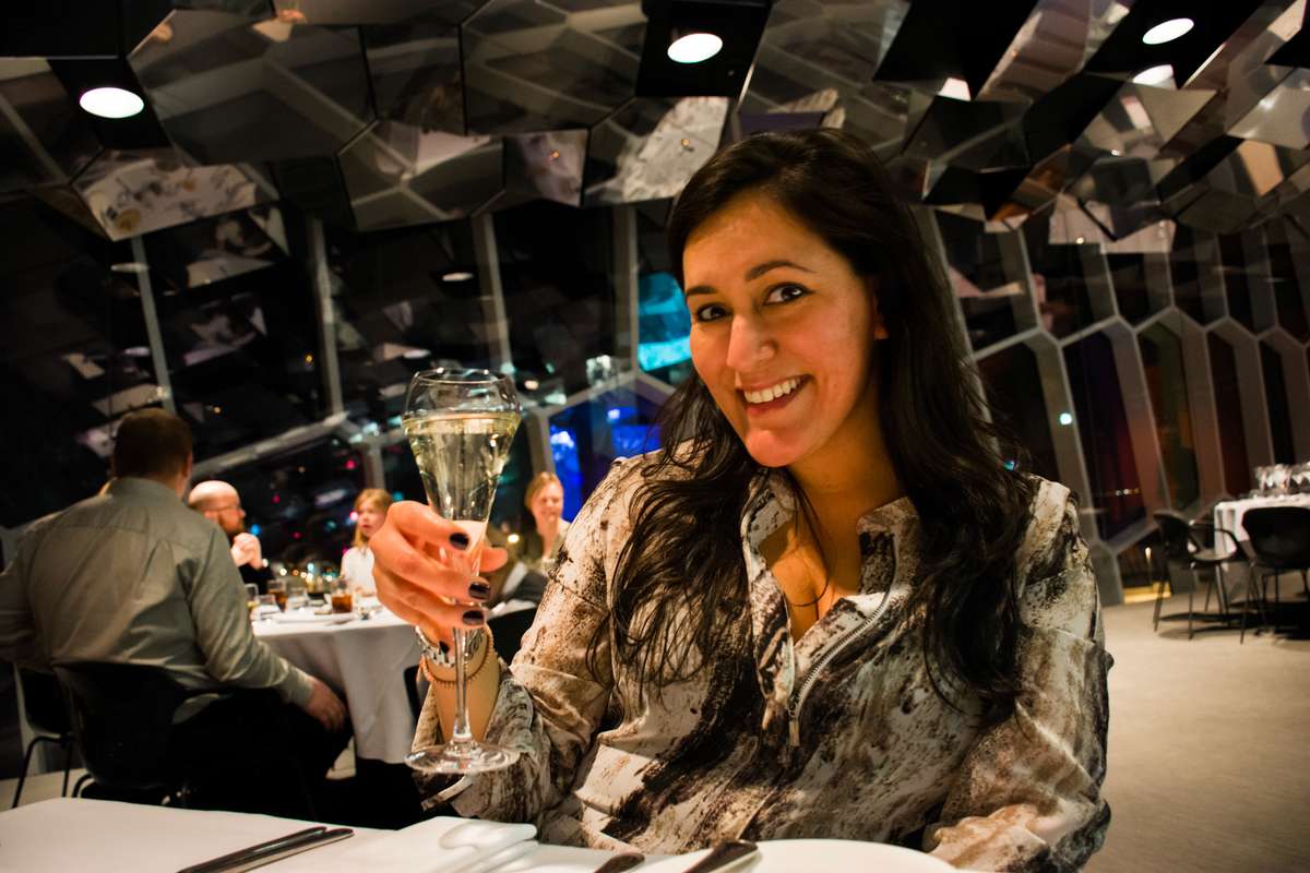 A smiling woman in a patterned shirt holds a glass of beer in a dimly lit bar with industrial decor and seating.