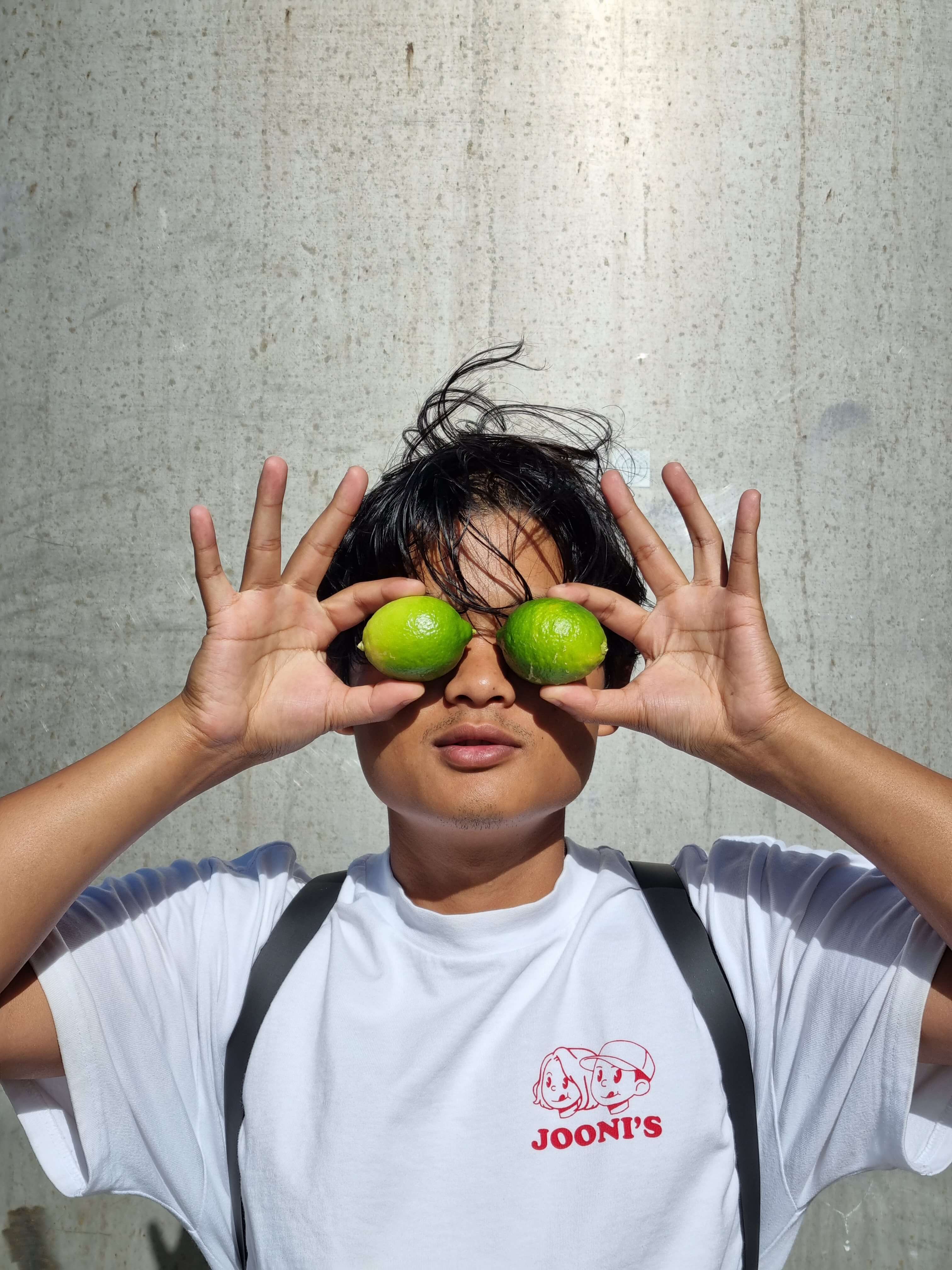 Young man with dark hair wearing a white tee shirt and backpack holding two limes up over his eyes