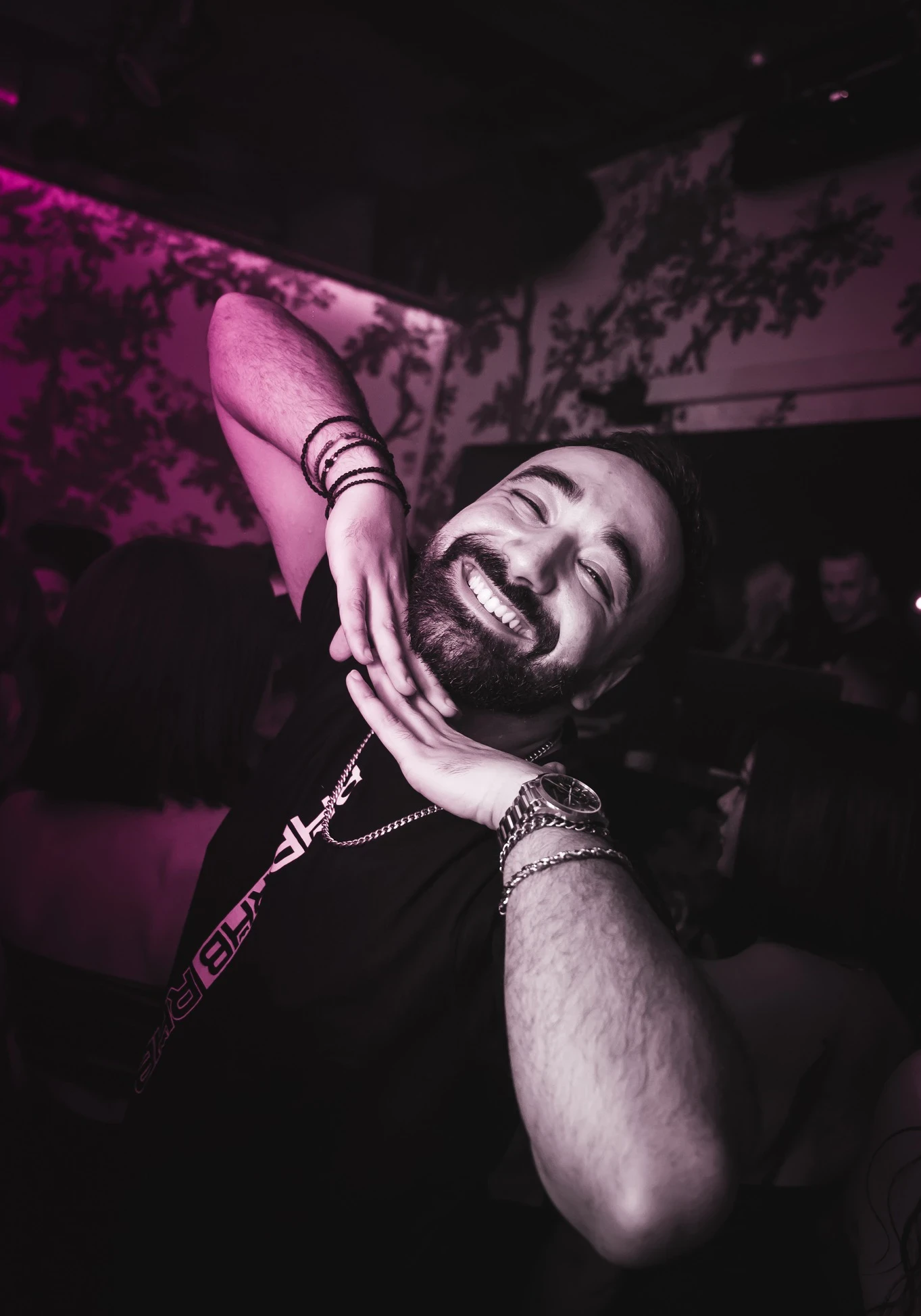 Liakakis, a man with a beard smiles widely, tilting his head and framing his face with his hands. He is wearing a black "RHB DRIP" t-shirt, necklaces, and bracelets at the party.