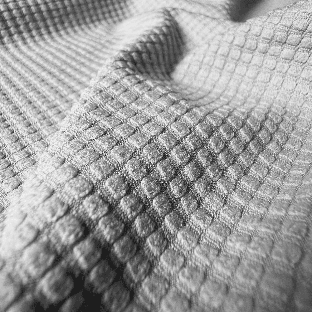 Macro close-up of a grey waffle-weave upholstery fabric used for ergonomic office chairs. The image showcases the intricate 3D honeycomb texture, highlighting the material's breathability, durability, and soft tactile feel
