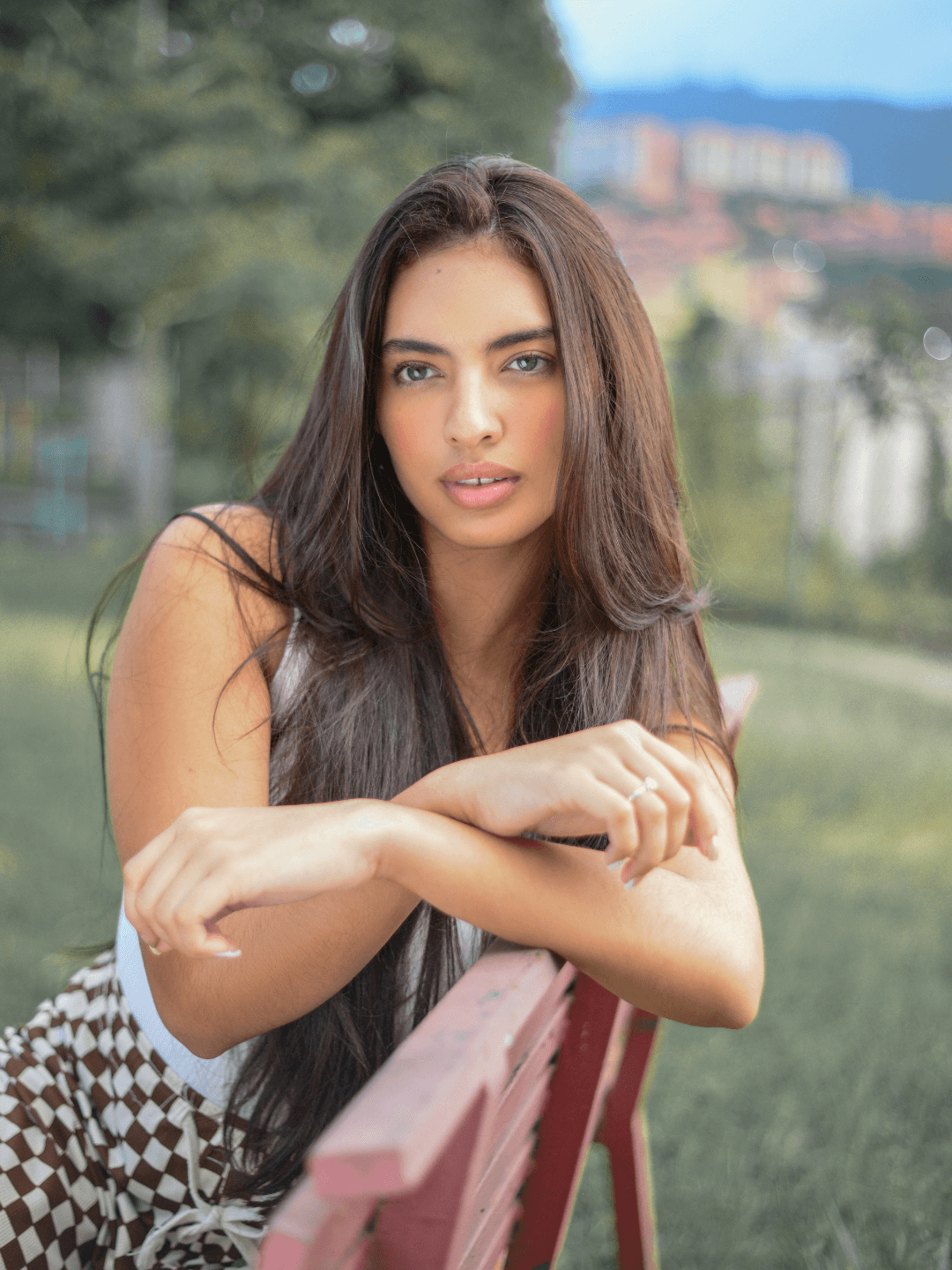 Portrait of a woman sitting on a bench in a park and resting her crossed arms on the back. She is looking at the camera