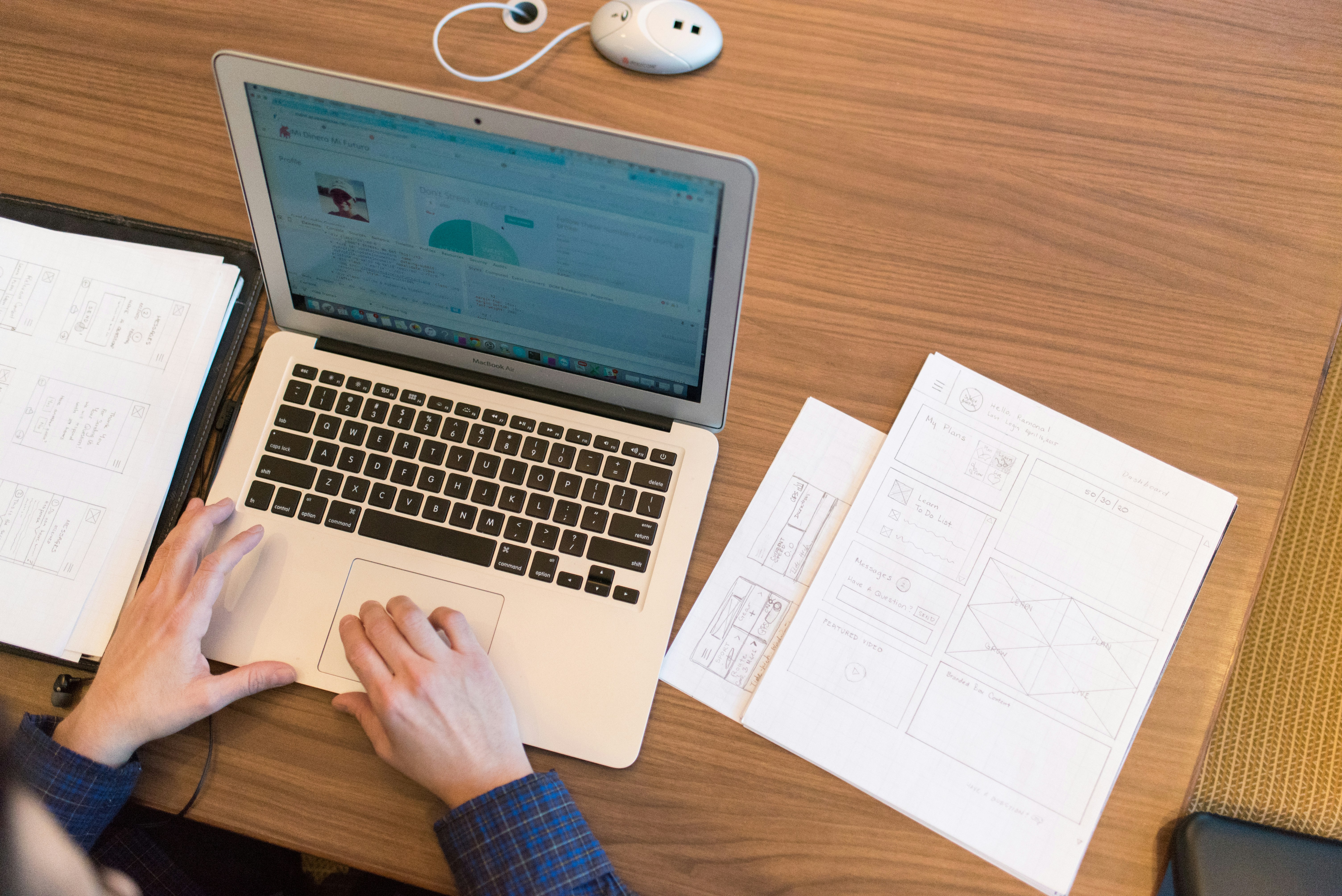 Overhead view of a person using a laptop beside printed wireframes and planning notes on a wooden desk.