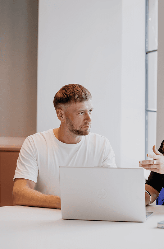 Man in white t-shirt sits at a table with a laptop, listening.