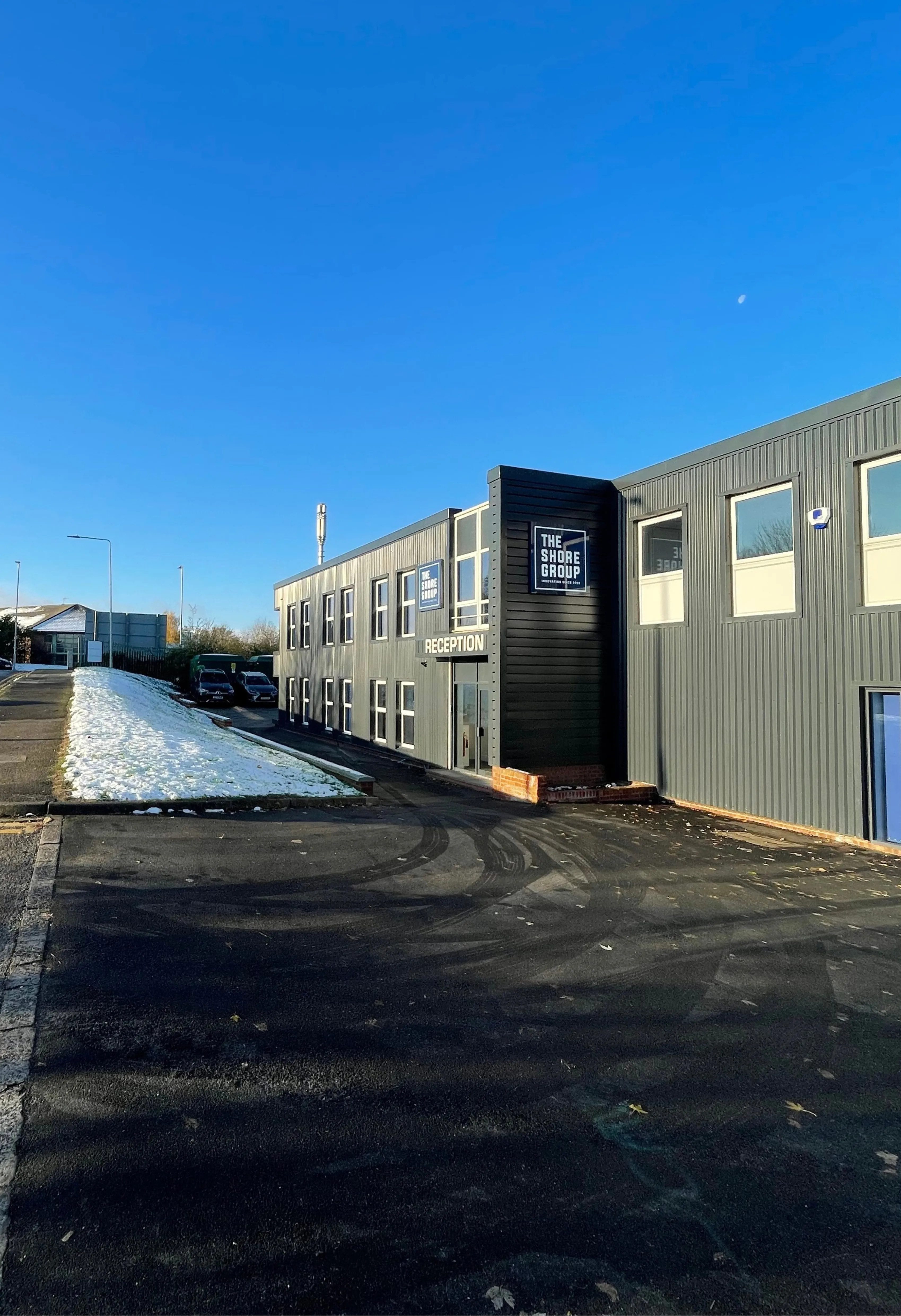 An industrial-style building with a modern gray facade and clear blue skies above, featuring a reception entrance and adjacent parking area, with a small snow-covered grassy section nearby.