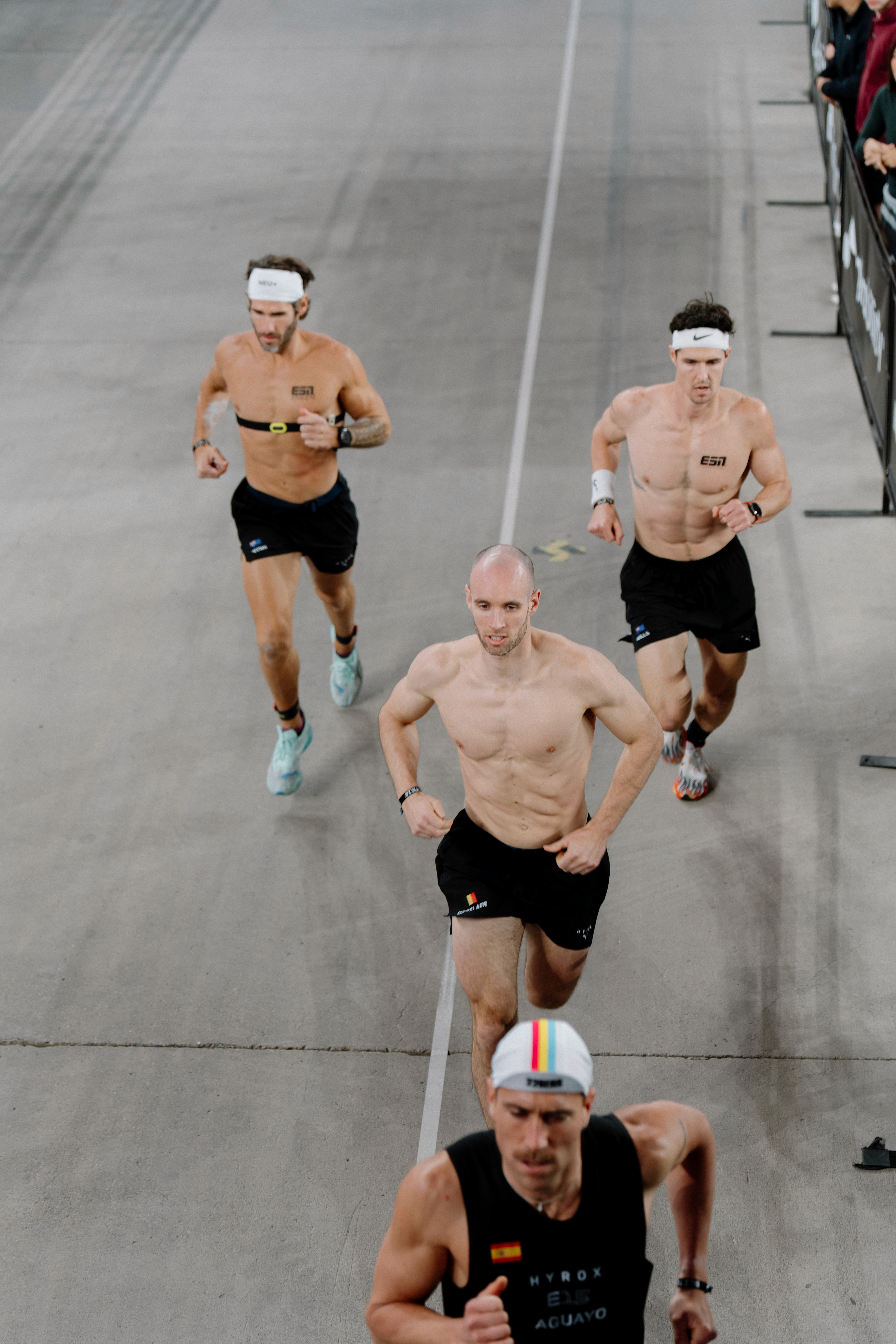 A dimly lit gym scene featuring a barbell rack and weights on the floor, with large windows in the background.