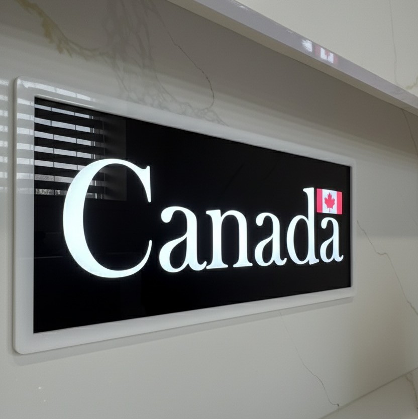 A sleek, rectangular interior sign featuring the Canada wordmark with illuminated white letters on a black background, framed in white and mounted on a marble wall in Calgary.