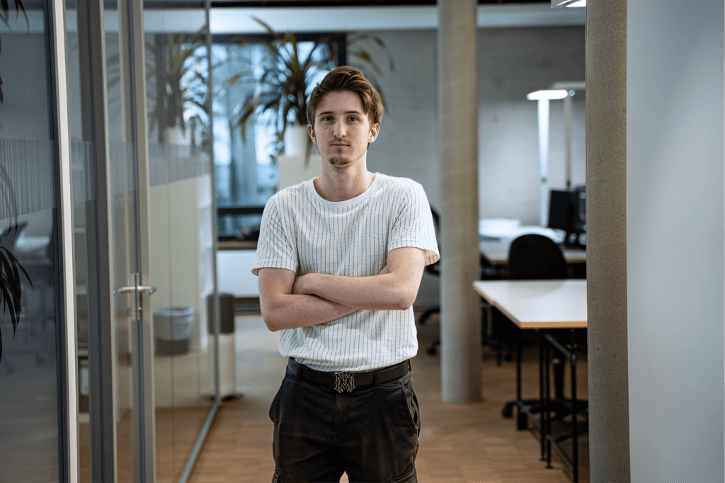 Junger Mann mit verschränkten Armen steht in einem modernen Büroflur der kba Architekten und Ingenieure GmbH; im Hintergrund sind Glaswände, Pflanzen und Arbeitsplätze zu sehen.