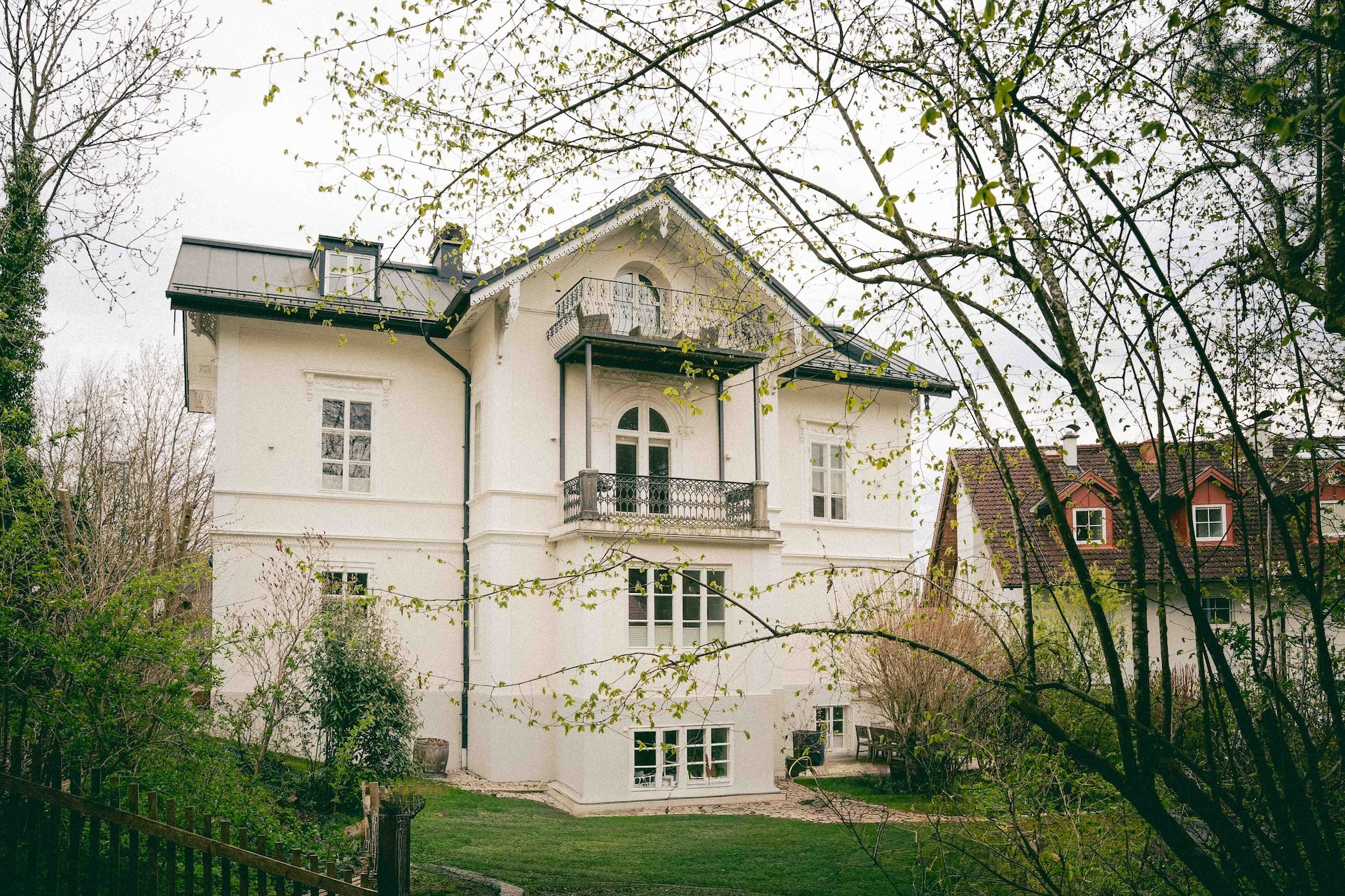 Weißes Haus mit Balkon und Rundbogenfenstern, von Büschen umgeben, mit angrenzendem Haus im Hintergrund.