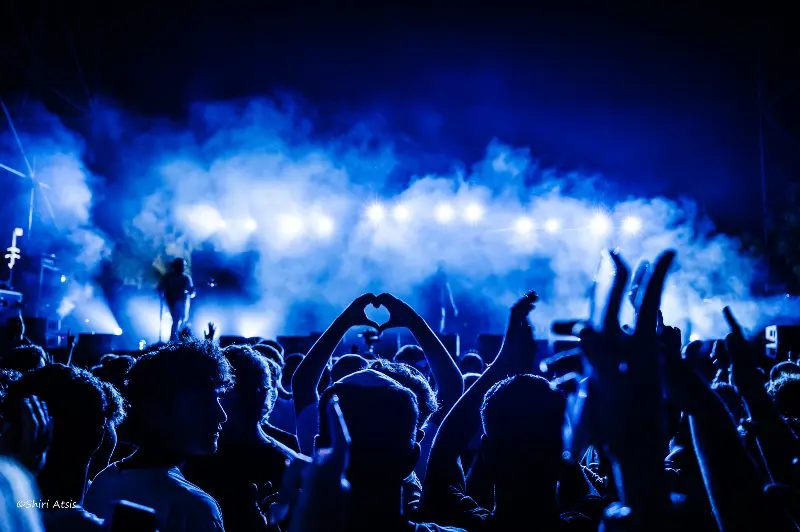 צילום אמנותי של קהל בהופעה חיה תחת תאורה כחולה עוצמתית, ידיים יוצרות צורת לב מול הבמה. צילום של הצלמת שירי עציץ Shiriz המתעד רגעי חיבור ורגש במוזיקה.