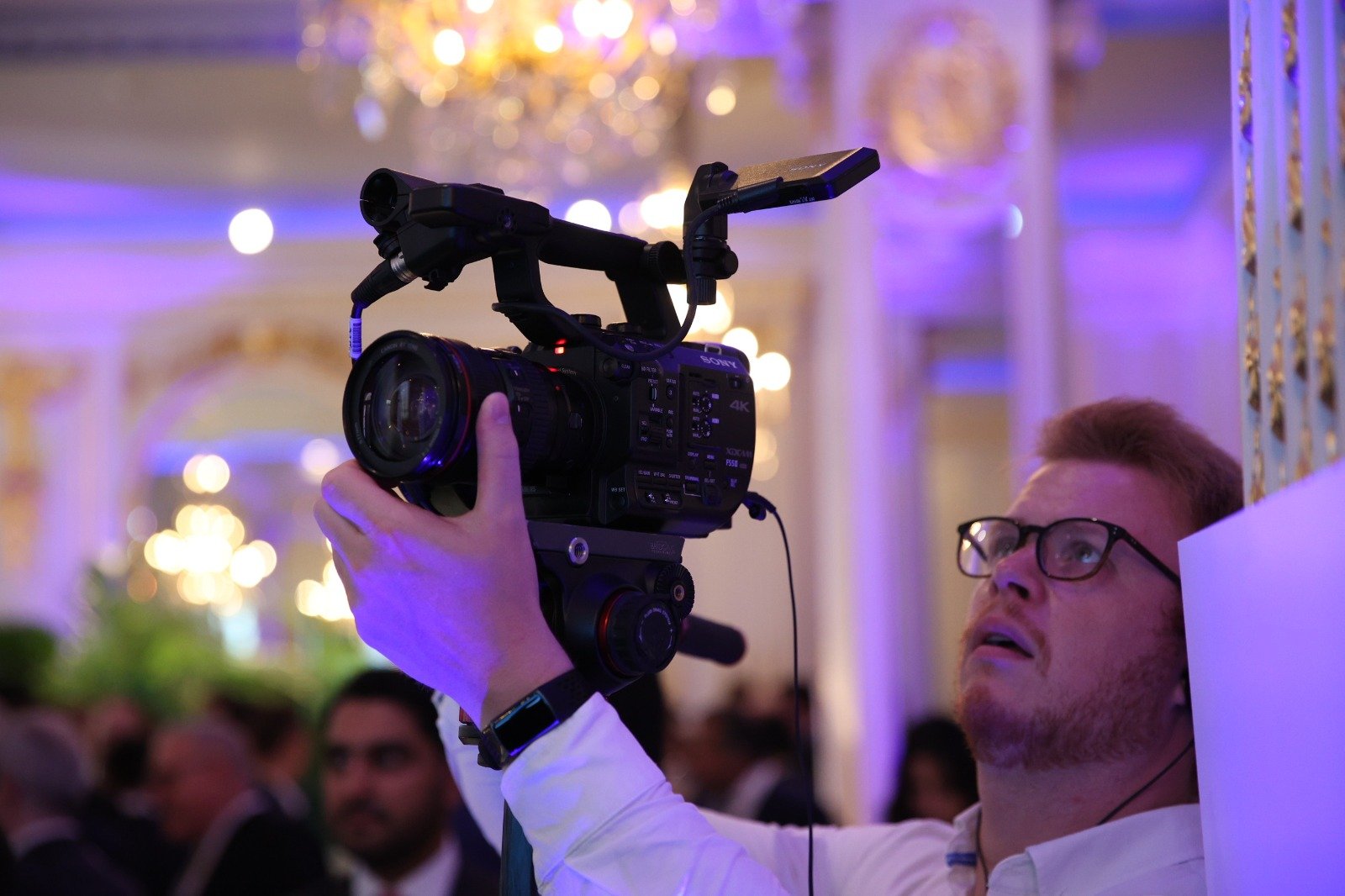 A videographer holding a camera at a crowded indoor event with purple lighting.