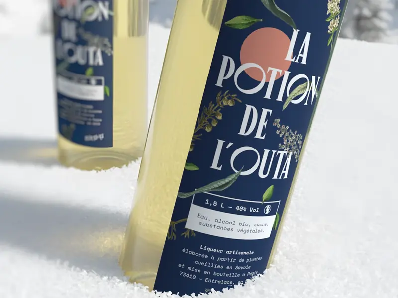 Bouteille de liqueur artisanale « La Potion de l’Outa » posée dans la neige, étiquette bleue illustrée avec typographie blanche et éléments végétaux.